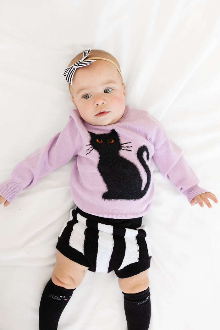 baby girl in purple cotton knit sweater with black fluffy cat design, black and white striped sweater bloomers and bow, and black cat knee highs for Halloween and witchy vibes