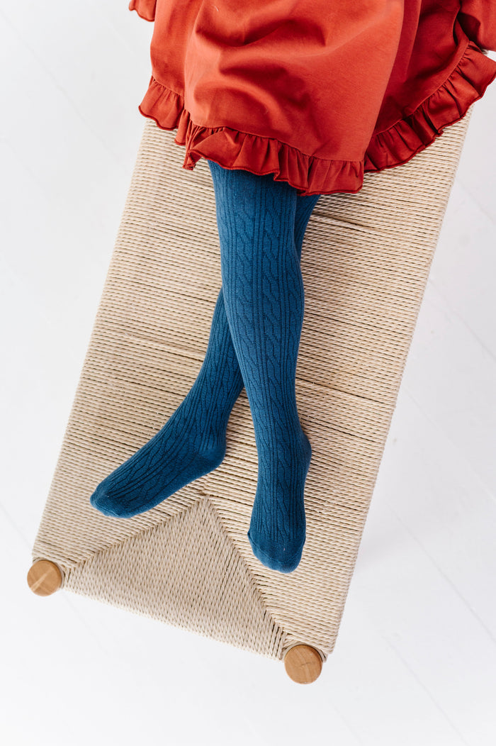 young girl sitting on bench in blue cable knit tights paired with rust ruffle hem twirl dress for fall
