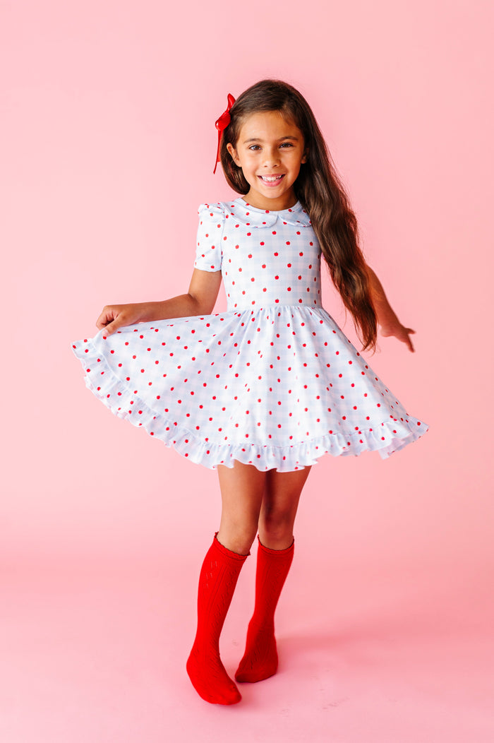 girl twirling in blue and white apple print gingham twirl dress paired with bright red pointelle knee socks and red satin hair bow