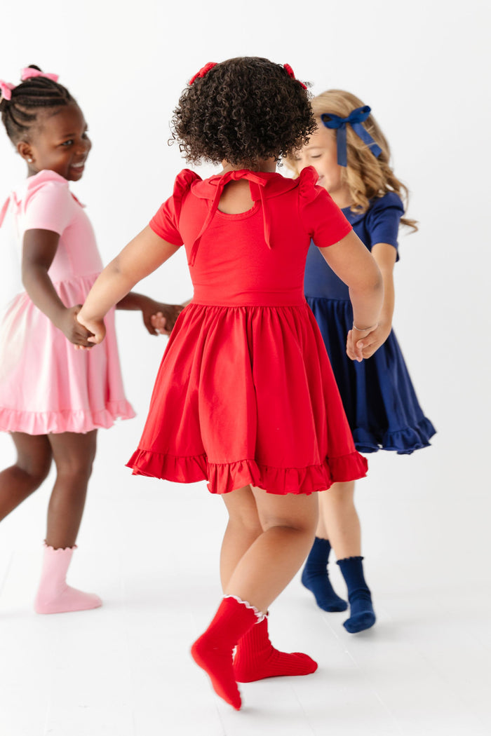 three girls playing ring around the rosies in solid uniform dresses and coordinating socks and bows with a back view of bright red twirl dress with tie back