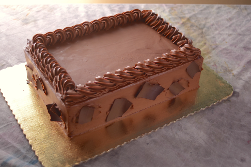 A rectangular cake covered in smooth chocolate frosting with a piped chocolate border around the top edge. The sides are decorated with large dark chocolate shards, and the cake is displayed on a gold cardboard base.