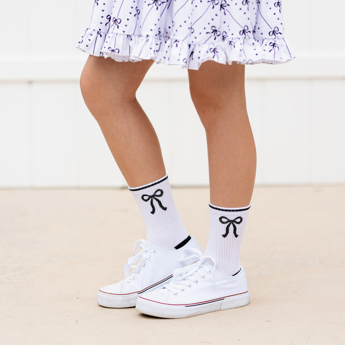 little girl in white crew socks with black bow design from 3-pack paired with matching white with black bow print twirl dress and white sneakers