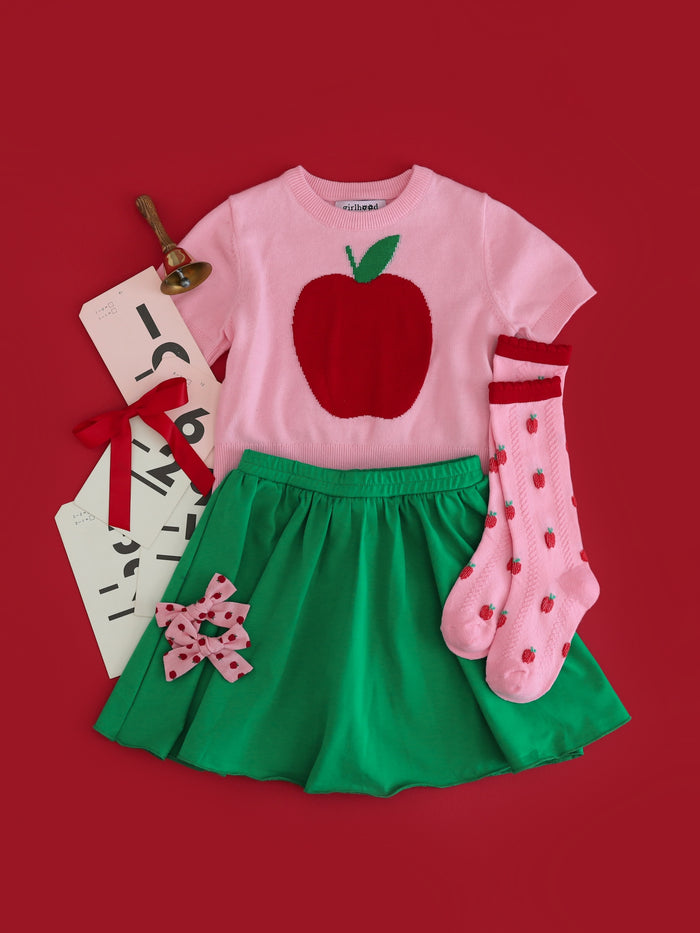 flatlay of back to school outfit including green cotton skort, pink with red apple short-sleeved sweater, pink knee highs with red apples and scallop trim and matching bows