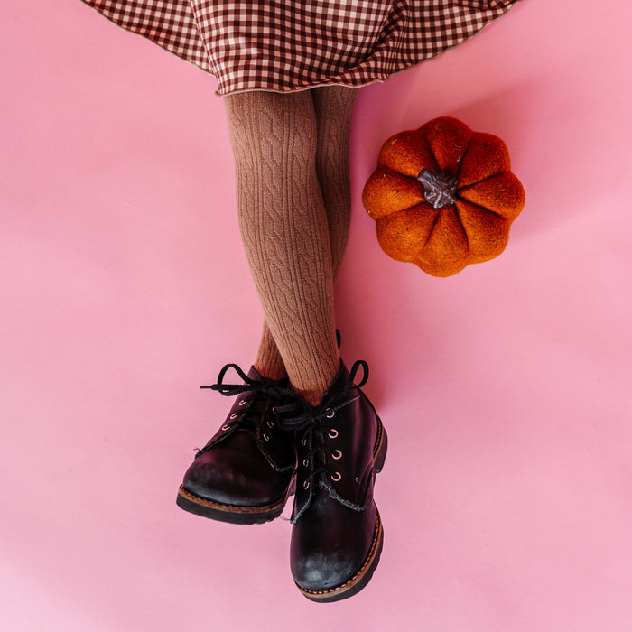 little girl sitting on pink backdrop by pumpkin decoration wearing light brown cable knit tights with brown gingham dress and black boots