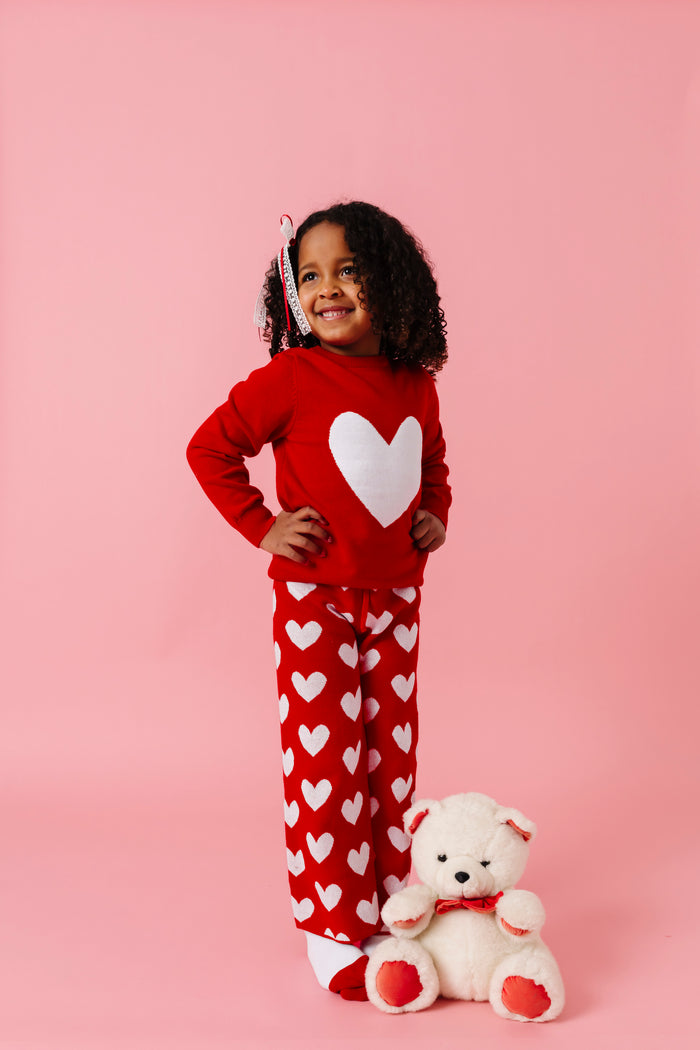 young girl with teddy bear wearing bright red sweater and pant set with white heart design