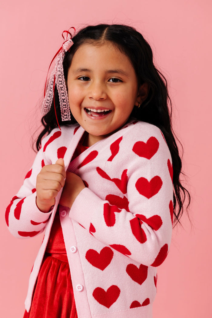little girl in soft pink cotton knit cardigan with buttons and red heart pattern paired with red velvet set and pink and red lace ribbon hair bow