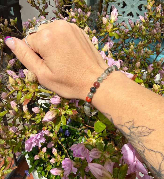 African bloodstone beaded bracelet with 6mm smooth round gemstones and sterling silver lobster claw clasp for courage and vitality on a models wrist with a flower background, front view.