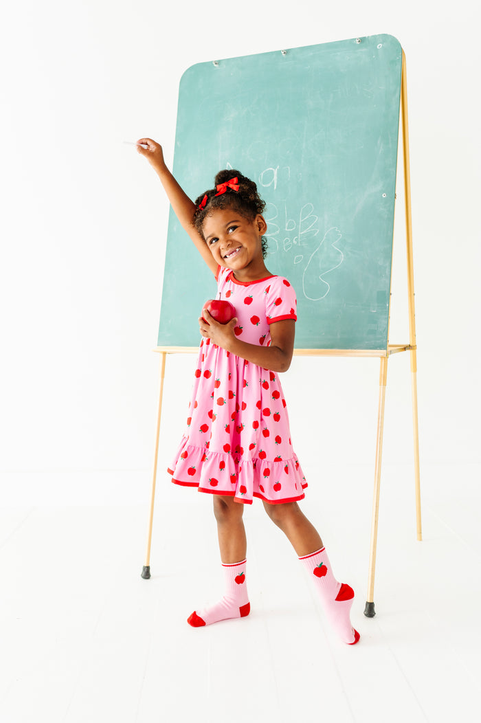 little girl in front of chalk board in pink dress with red trim and red apple print paired with matching pink apple crew socks and red satin pigtail bows