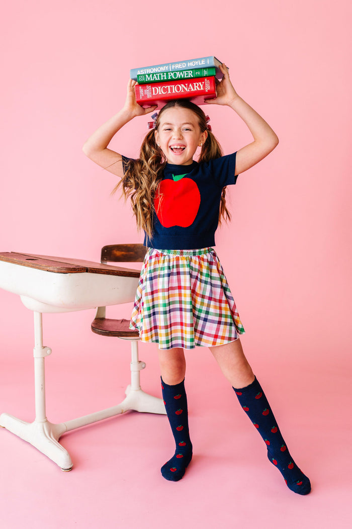girl wearing back to school outfit with red apple top, apple knee high socks and rainbow gingham skort