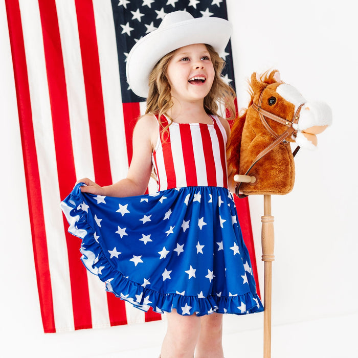 little girl with stick horse in front of American flag wearing flag print sleeveless twirl dress with white cowgirl hat and boots