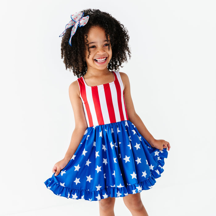 little girl in stars and stripes American flag sleeveless twirl dress and coordinating sheer hair bow with blue trim and red dots