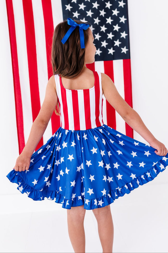 little girl in front of American flag wearing matching stars and stripes twirl dress and blue satin hair bow