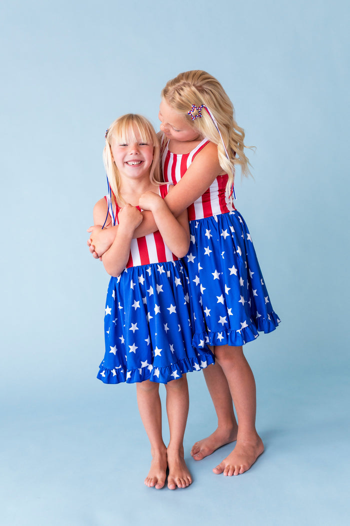 sisters in matching sleeveless American flag twirl dresses and matching red, white and blye braises star clips with ribbon streamers for the 4th of July