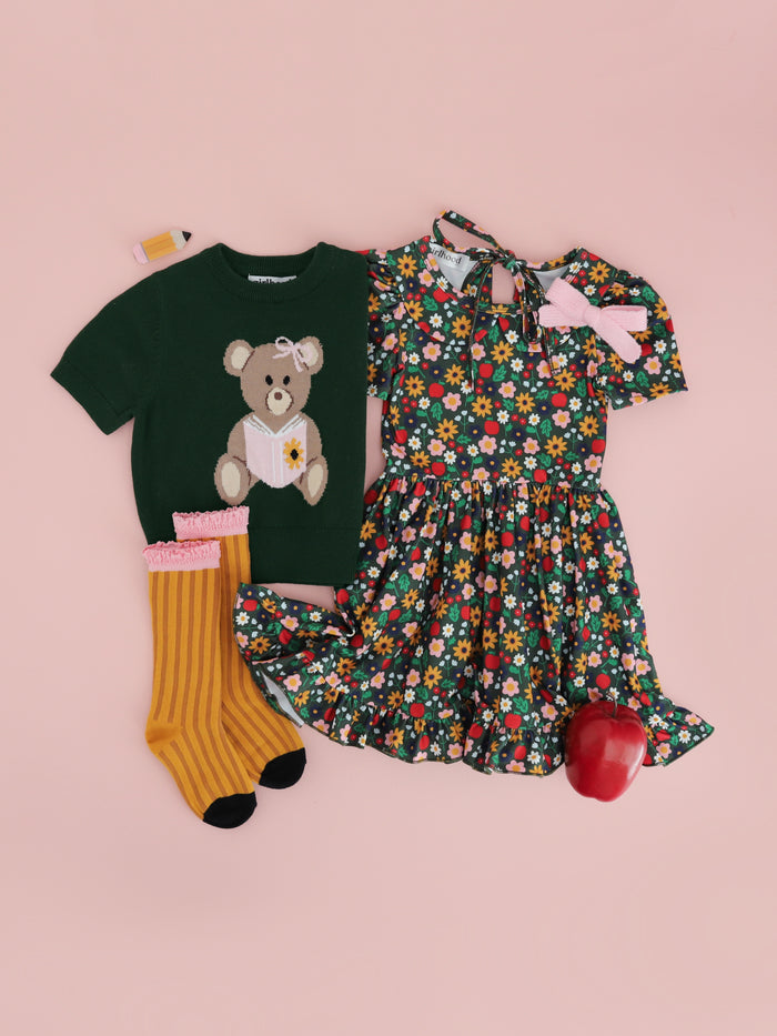 girls' styled back to school outfit with green floral and apple print twirl dress, coordinating green short sleeve knit sweater with bear design and matching pencil socks and clip