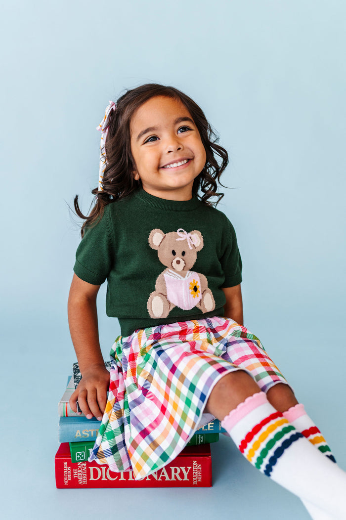 little girl sitting on books in green short sleeve knit sweater with bear reading book design paired with primary gingham skort and matching knee socks