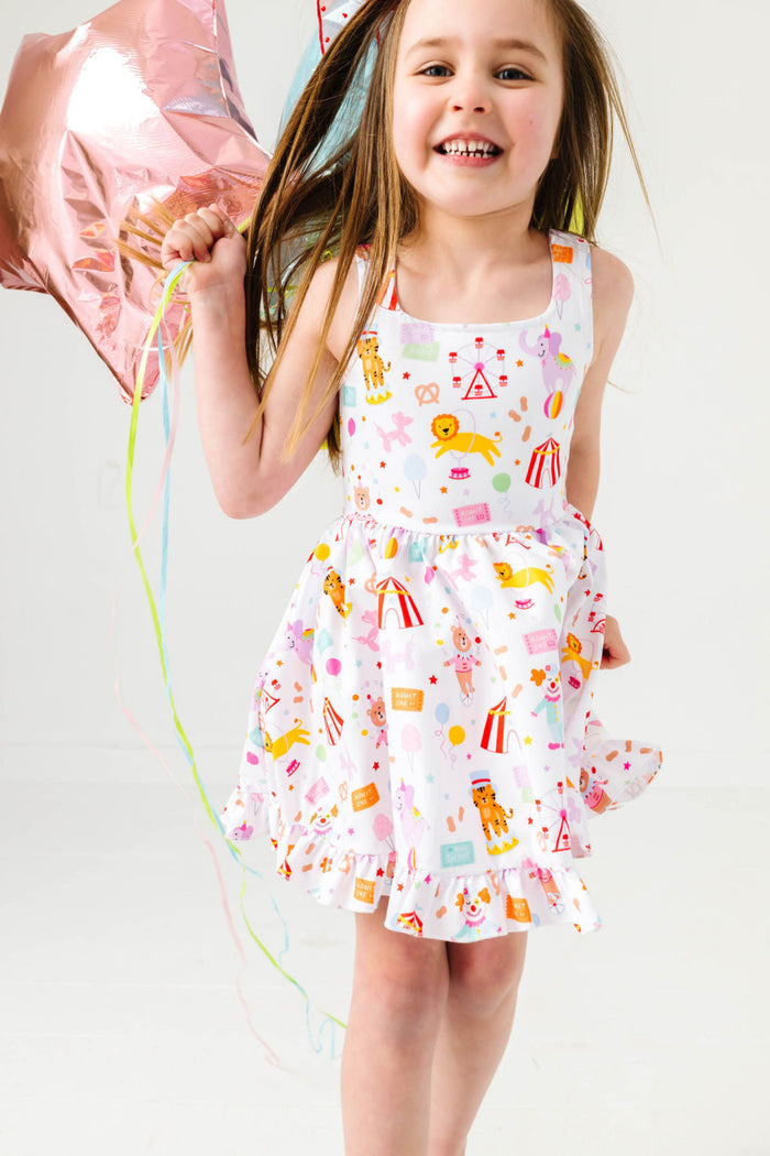 little girl jumping with balloon in sleeveless summer twirl dress in white featuring colorful circus themed print