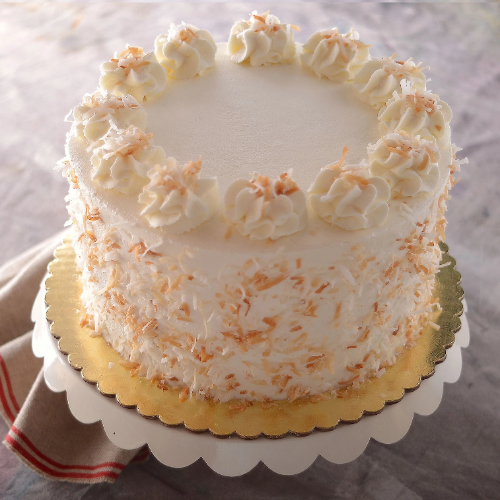 Round cake with white frosting, toasted coconut topping and sides, on gold board and scalloped white stand.