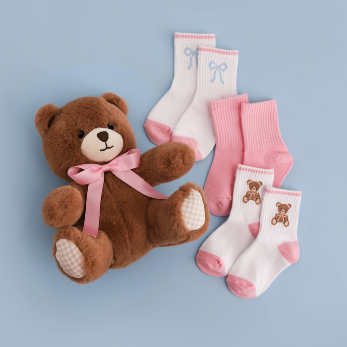 flat lay of girls' teddy and bow themed three pack of crew socks beside stuffed bear toy: white with blue bows, solid pink, white with teddy design