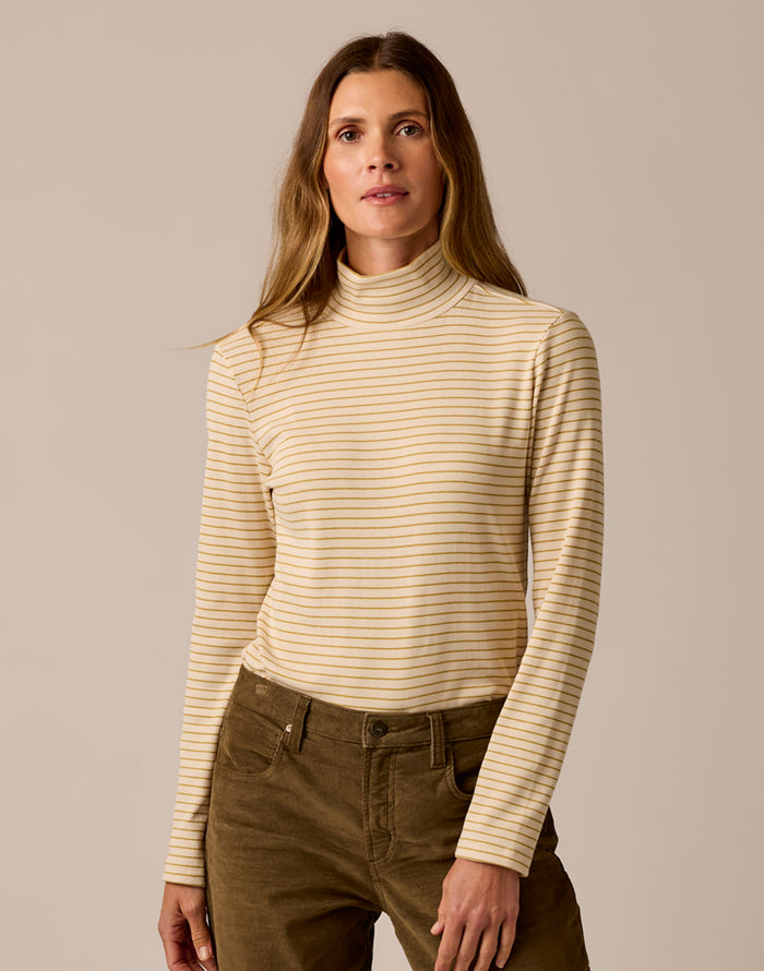 Woman wearing a beige striped turtleneck sweater and brown pants against a beige background