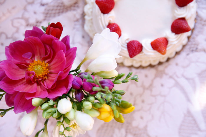 Colorful flowers in foreground with a white-frosted cake topped with strawberries in the background on lace table.