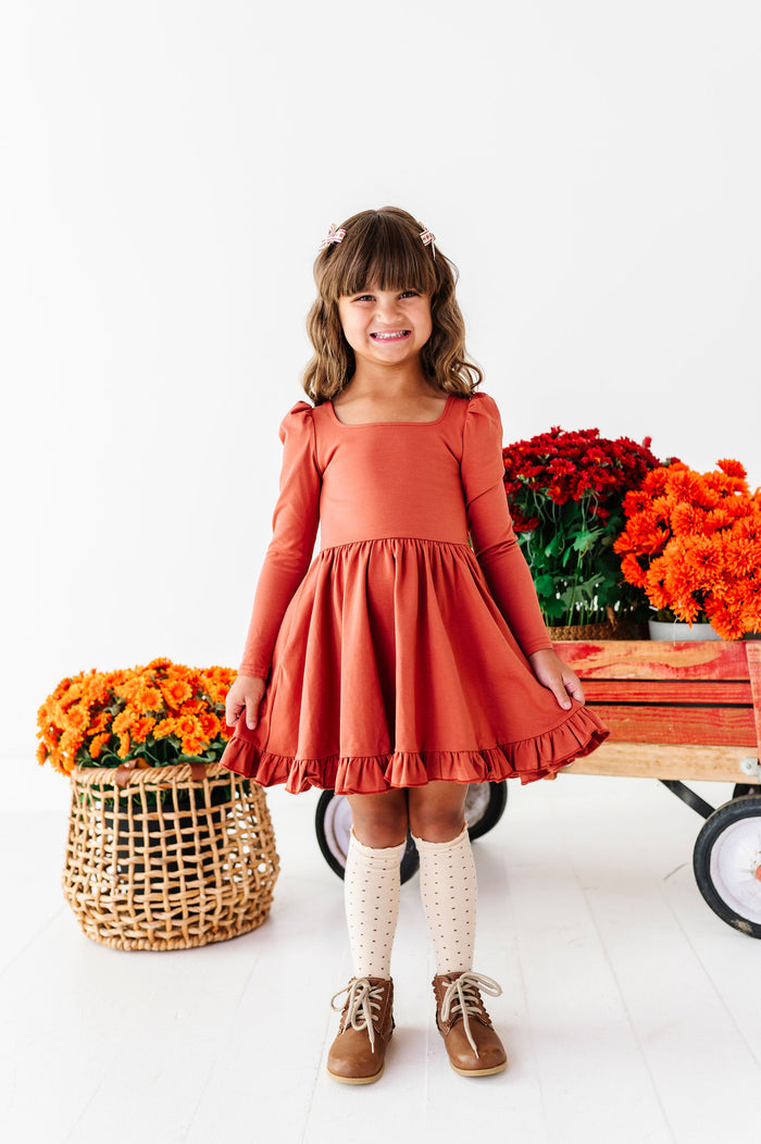 young girl in front of wagon filled with fall flowers wearing rust long sleeve cotton blend twirl dress with square neckline and coordinating cream dotted knee socks