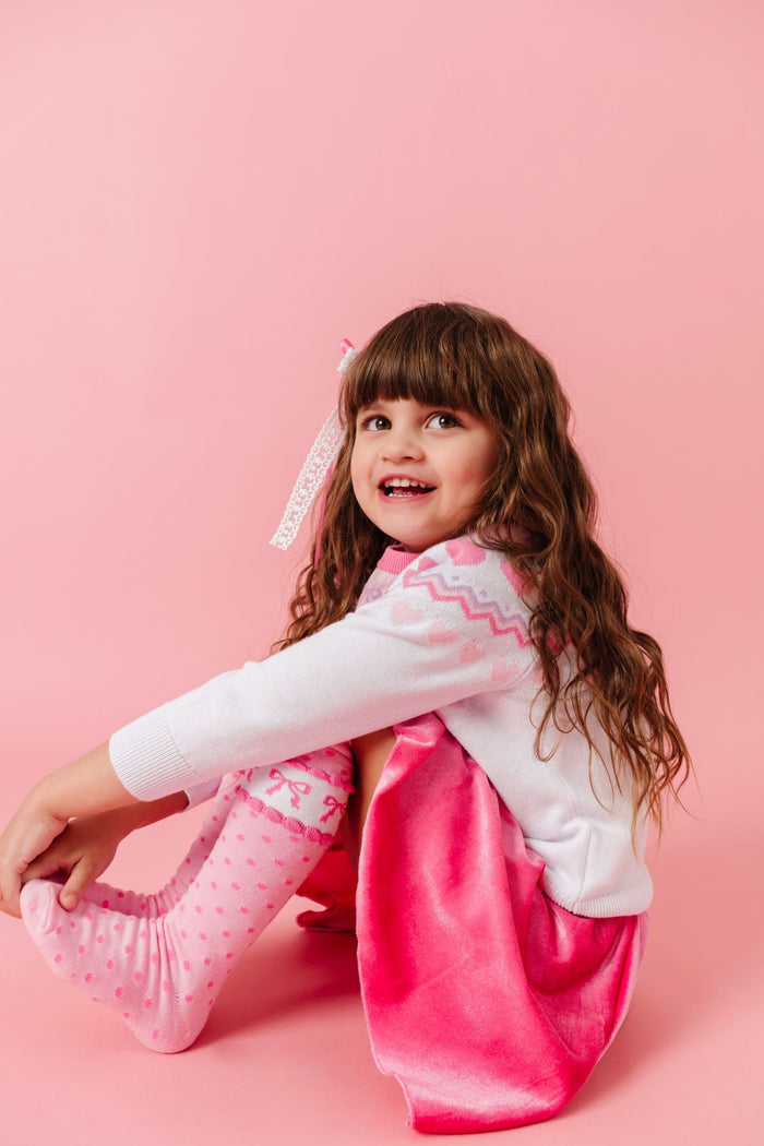 little girl sitting in valentines fair isle sweater, pink velvet skort and matching pink bow knee socks