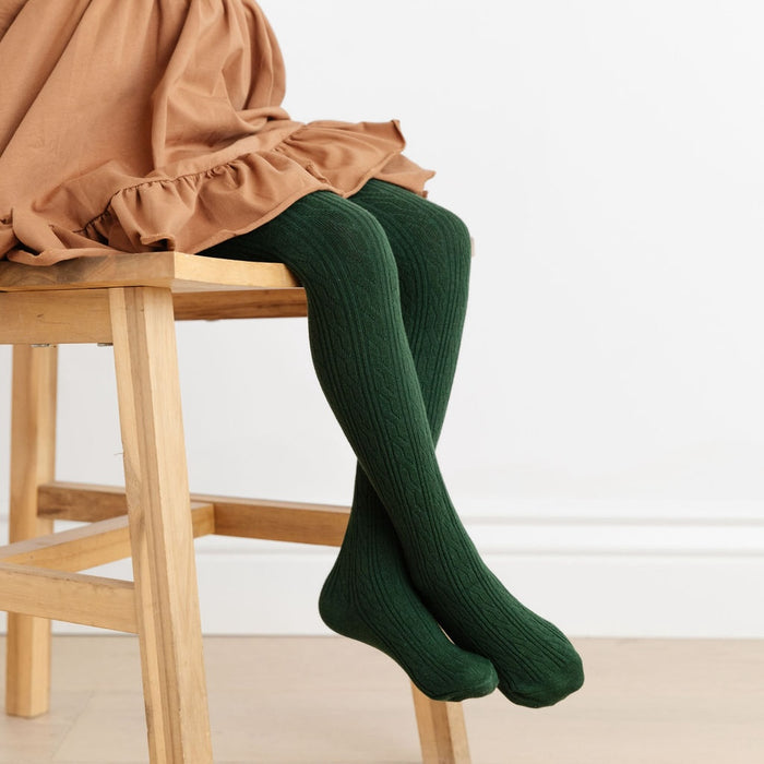 little girl on wooden stool wearing tan dress and forest green cable knit tights by little stocking co.