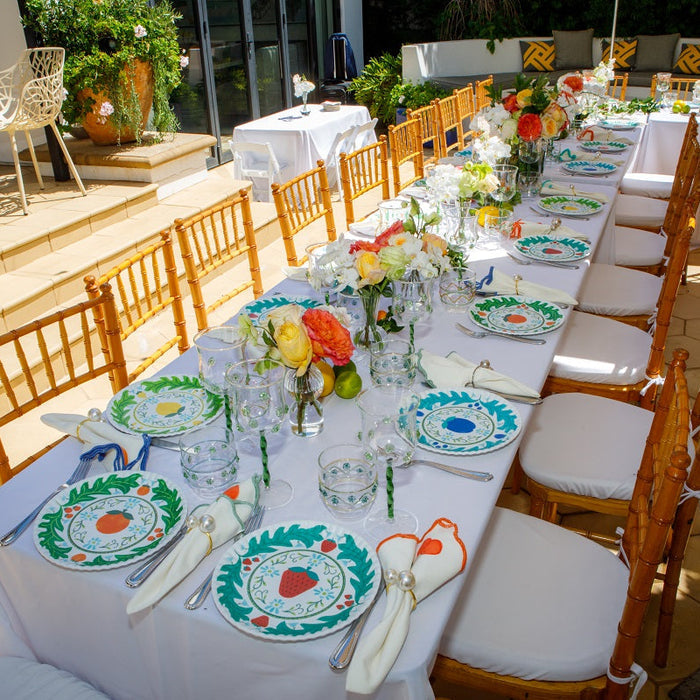 An outdoor event table setup with a long white tablecloth, matching round plates with teal and orange floral borders, clear glassware, folded white napkins, and wooden chairs on a sunlit patio.