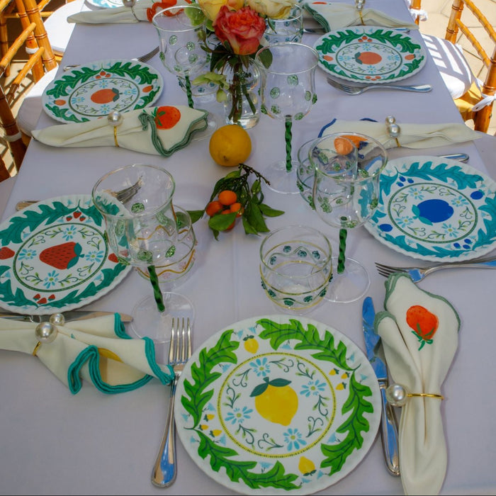 A long outdoor dining table set with white tablecloths, hand-painted green and blue ceramic plates with lemon and strawberry motifs, matching napkins, and floral centerpieces.