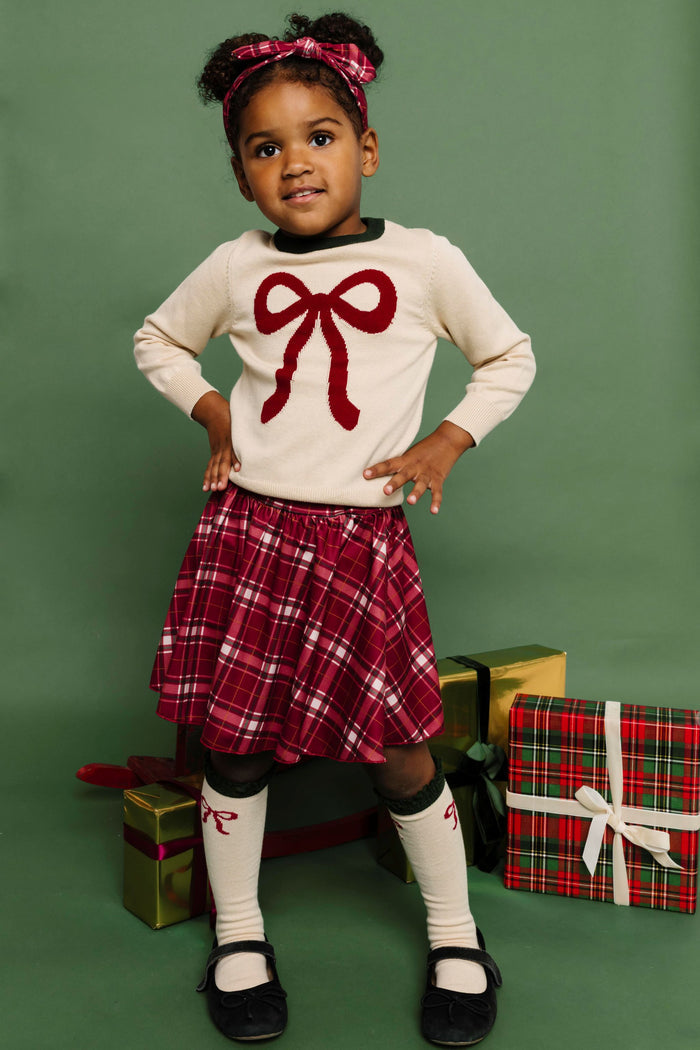 little girl with Christmas gifts in matching cream sweater and knee socks with crimson bow design paired with crimson plaid skirt and headband