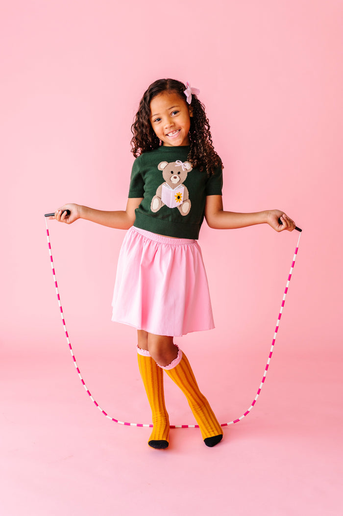 girl with jump rope wearing green school sweater with bear reading book paired with matching pink skort and pencul knee socks