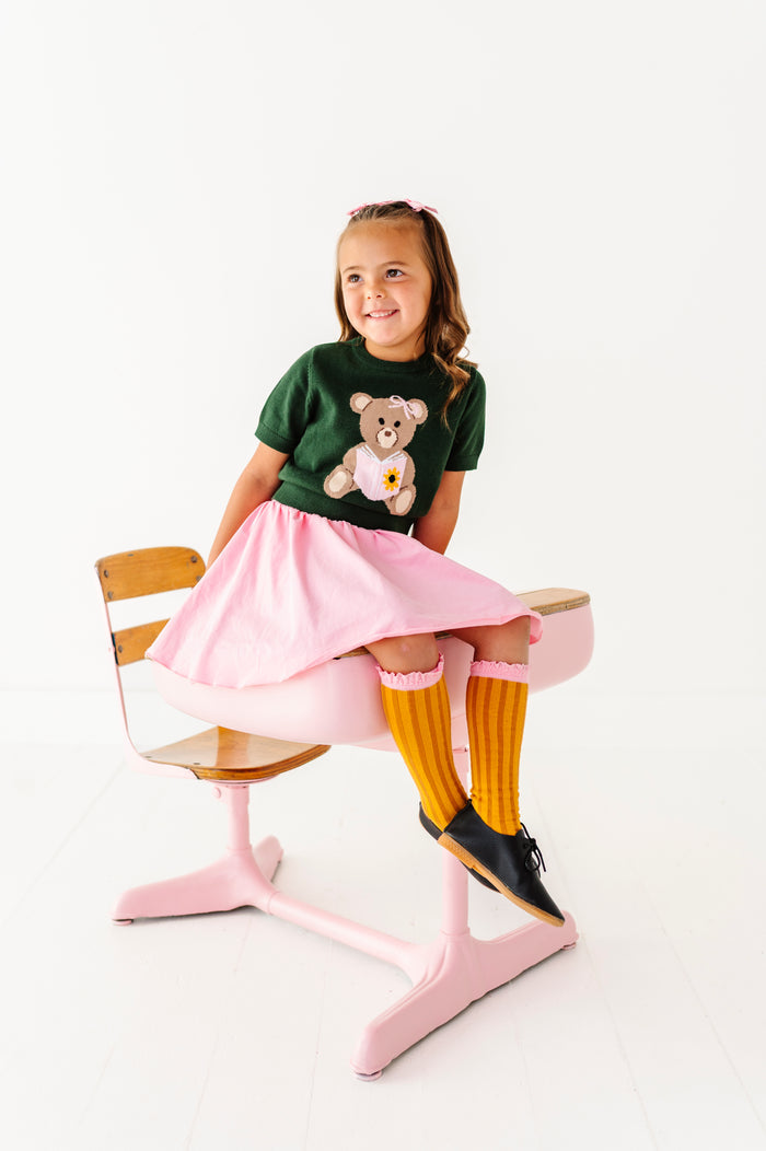 girl sitting on desk in green short sleeve bear sweater, pink skort and pencil themed knee socks for back to school