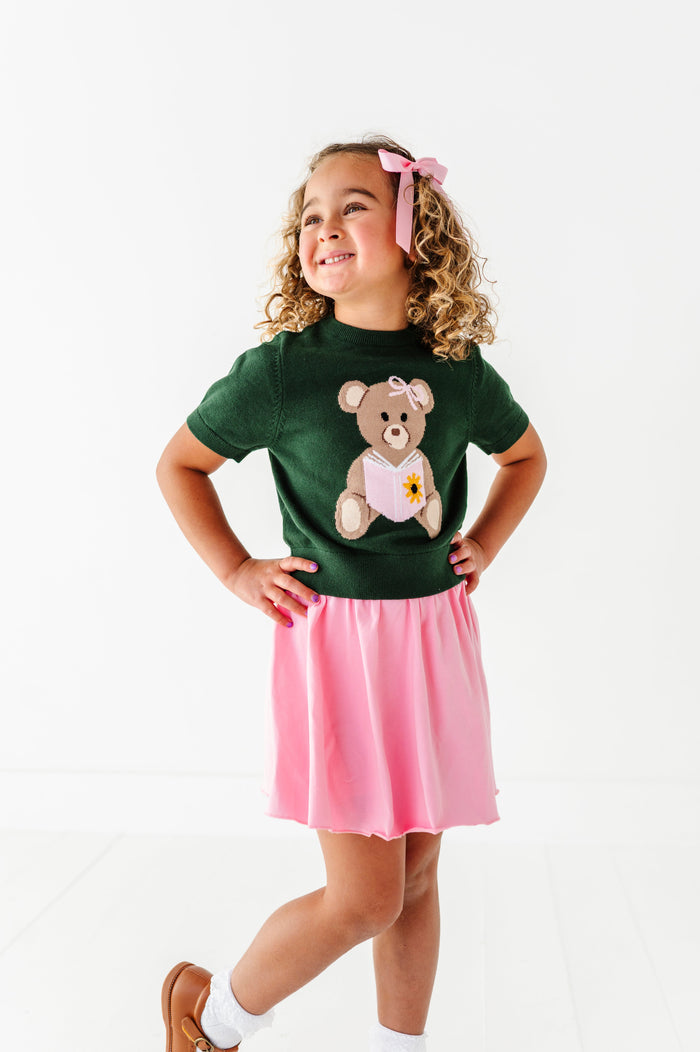 young girl in forest green short sleeve sweater with bear holding a book design and matching light pink twirl skort and hair bow