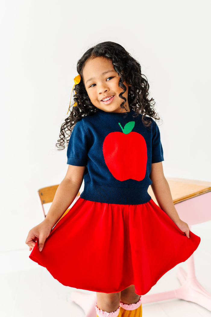young girl in front of school desk in navy short sleeve apple sweater paired with matching bright red skort