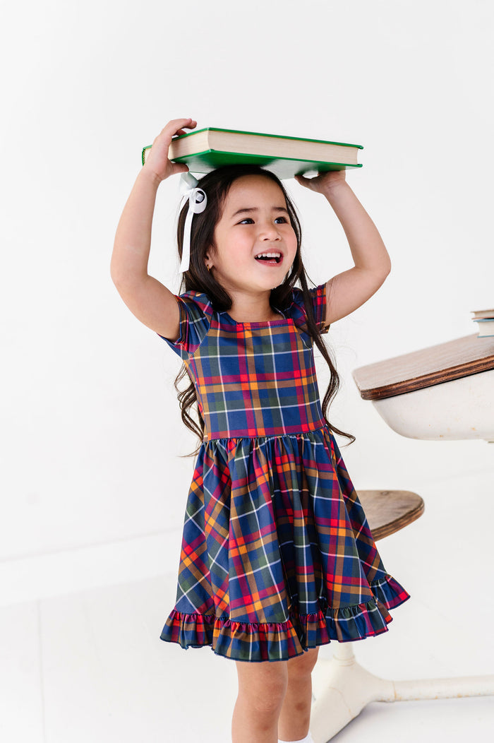 girl wearing navy and primary color plaid dress and holding a book for back to school