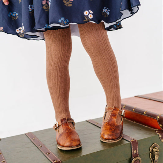 little girl standing on vintage suitcase in light brown cable knit tights paired with navy blue floral twirl dress and brown mary janes