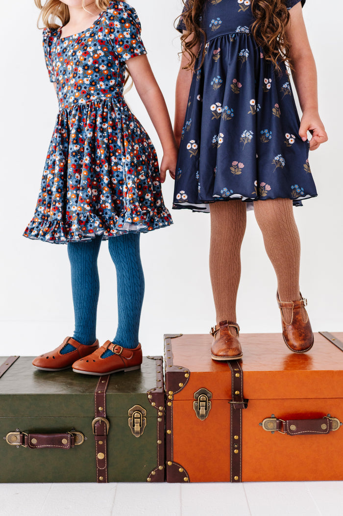 two models standing on vintage luggage in coordinating blue floral twirl dresses paired with matching blue and light brown cable knit tights