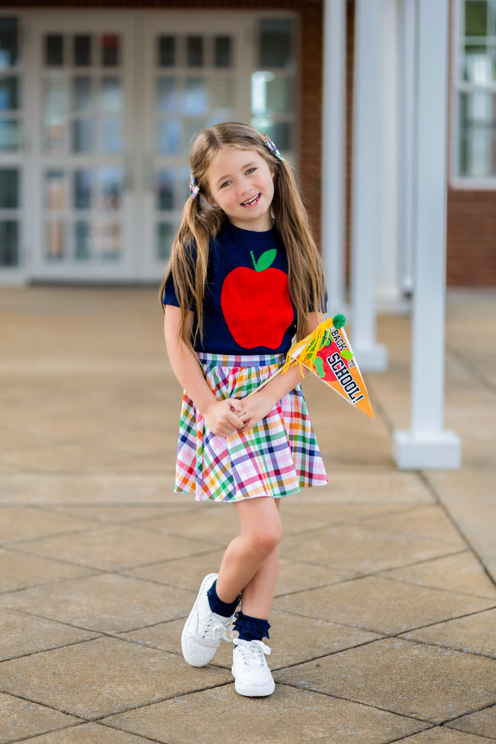young girl with back-to-school flag wearing navy short sleeve knit apple sweater and coordinating gingham skort in primary colors