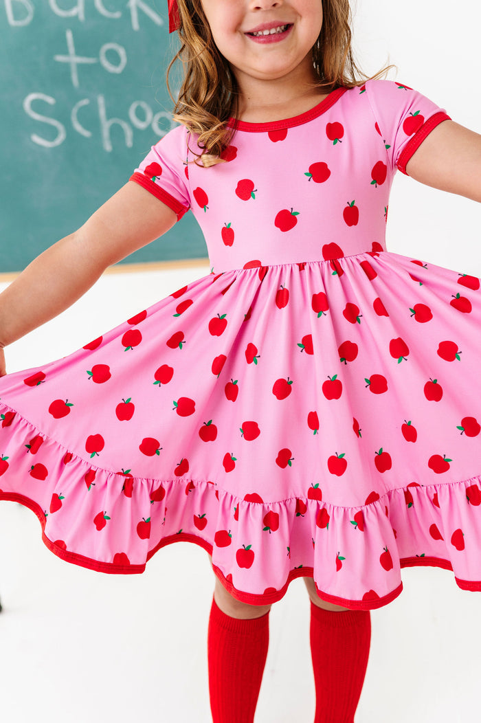 Girl wearing a pink dress with red apple pattern in front of a chalkboard.