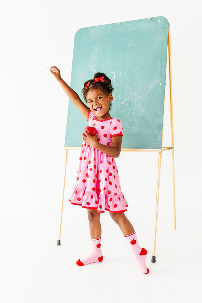 little girl raising hand in pink twirl dress with red trim and red apple design paired with matching apple crew socks