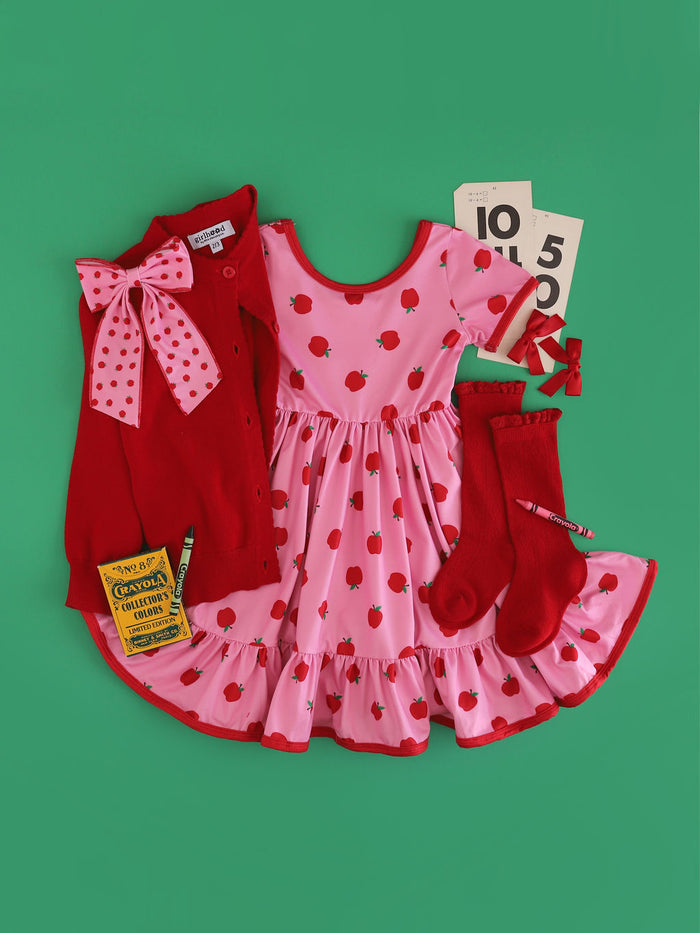 girl's pink apple print twirl dress with red trim paired with matching apple print bow, bright red cardigan and pointelle knee socks