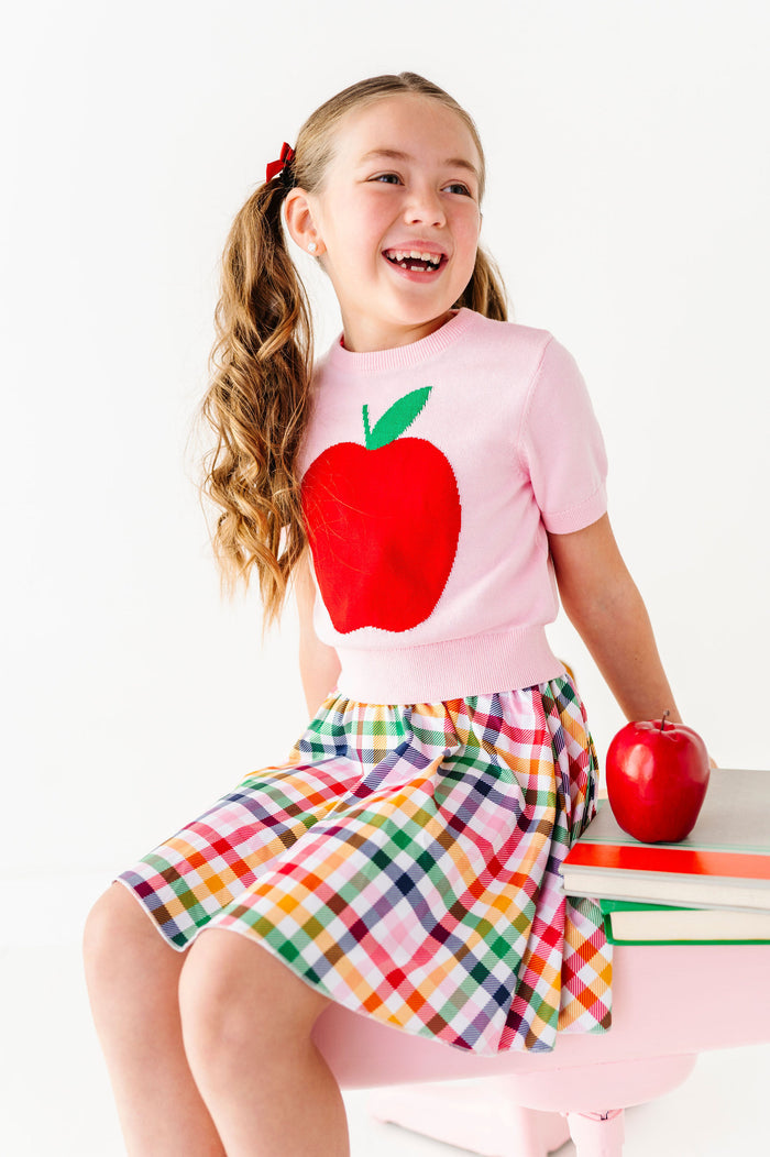 girl wearing pink top with big red apple and rainbow plaid skort for back to school