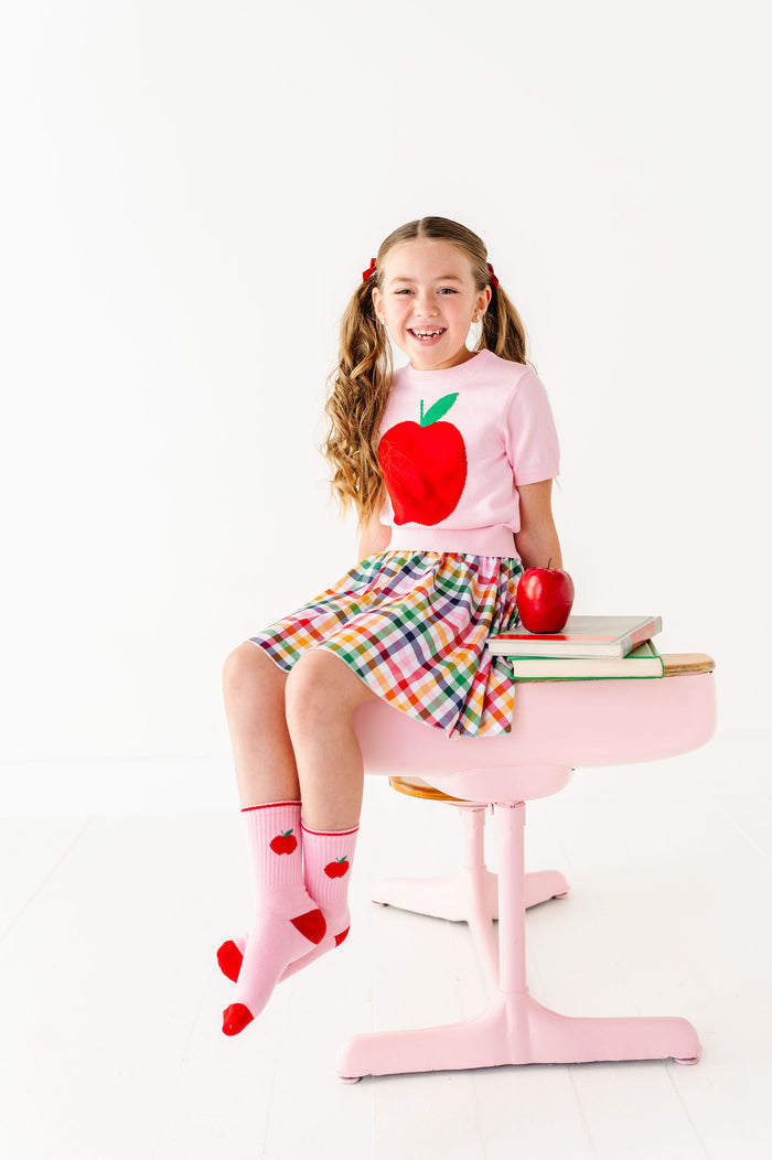 girl sitting on a pink chair wearing a pink shirt with an apple design, plaid skirt, and pink socks with apple pattern.