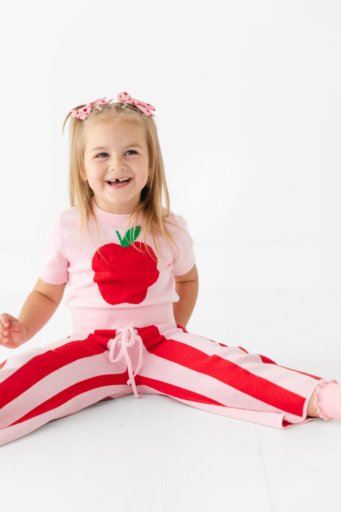 little girl sitting in red and pink striped cotton sweater pants and matching apple design short sleeve sweater with apple print pigtail bows