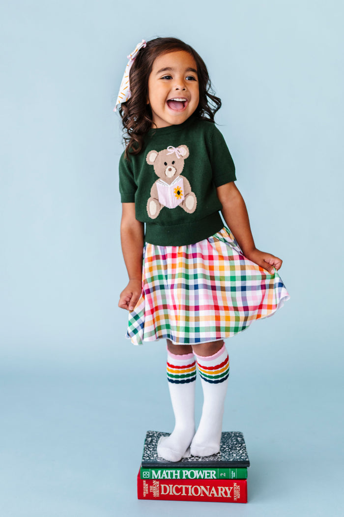 little girl standing on books in green short sleeve knit sweater with bear reading book design paired with primary gingham skort and matching striped knee socks