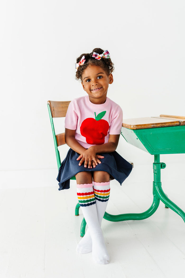 girl sitting at a desk wearing a shirt with an apple design and rainbow stripe knee high socks