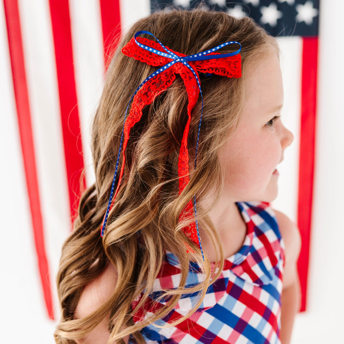 girl in front of American flag wearing red, white and blue gingham twirl dress and red lace with blue star ribbon hair bow
