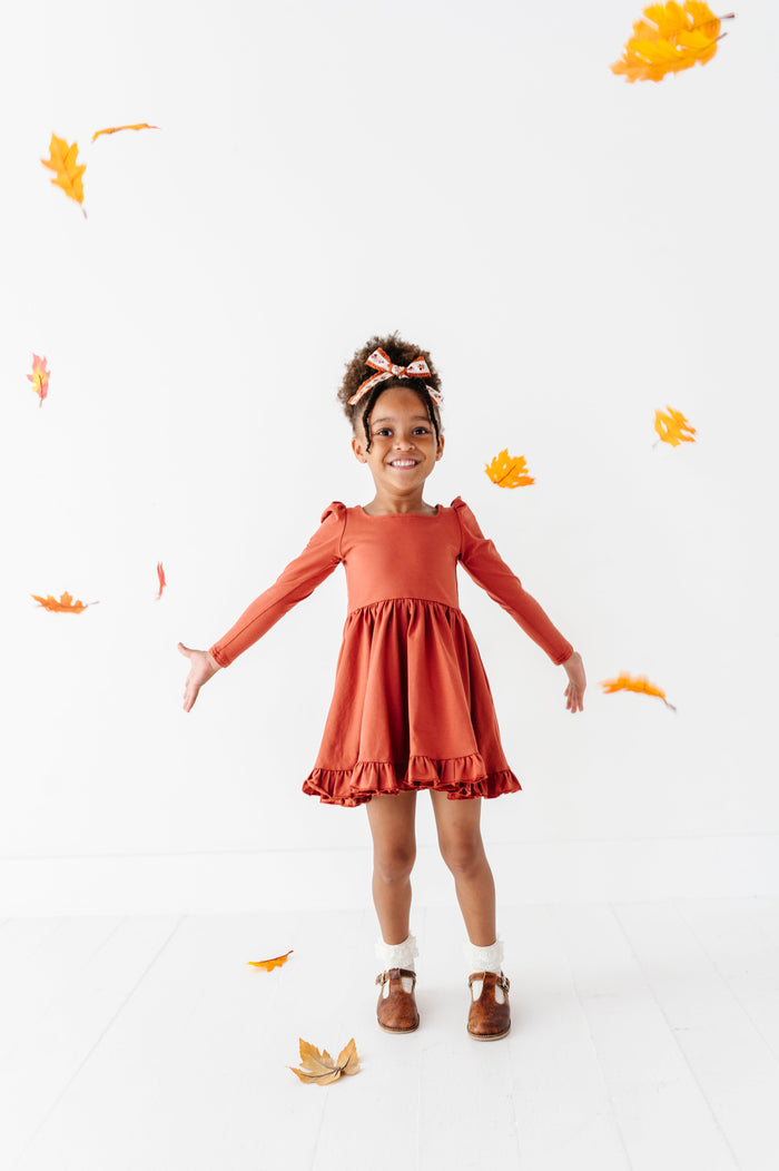 little girl throwing fall leaves in rust long sleeve twirl dress paired with ivory lace bobby socks and rust floral hair bow