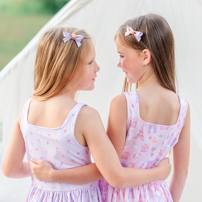 Two young girls in matching dresses with hair clips, standing close together outdoors.