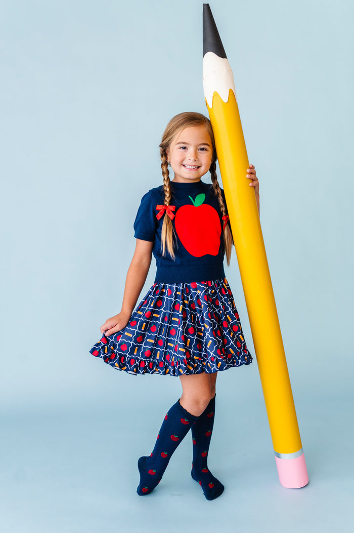 girl holding giant pencil prop in navy apple sweater with matching navy pencil and apple print twirl dress and navy apple socks